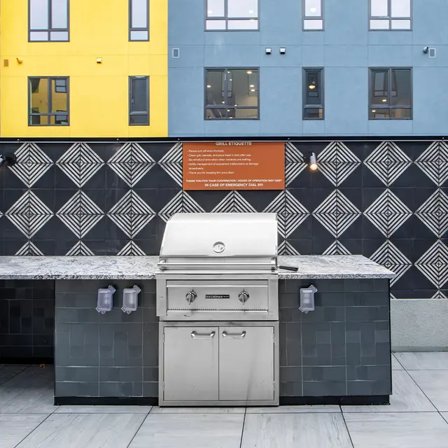 An outdoor kitchen area featuring a stainless steel grill, surrounded by dark tiled countertops and a decorative backsplash with a geometric pattern, set against a backdrop of colorful apartment buildings.