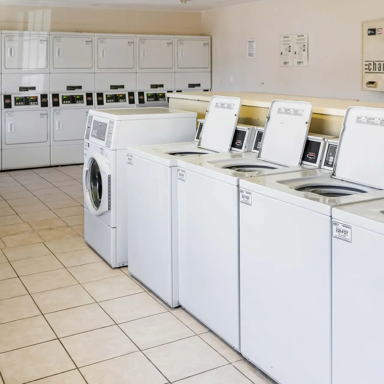 Haltere at Reserve - A clean and modern laundry room featuring multiple washing machines and a row of dryers along one side. There are empty tables and chairs for customers to use while washing clothes, and shelves for storage in the background.