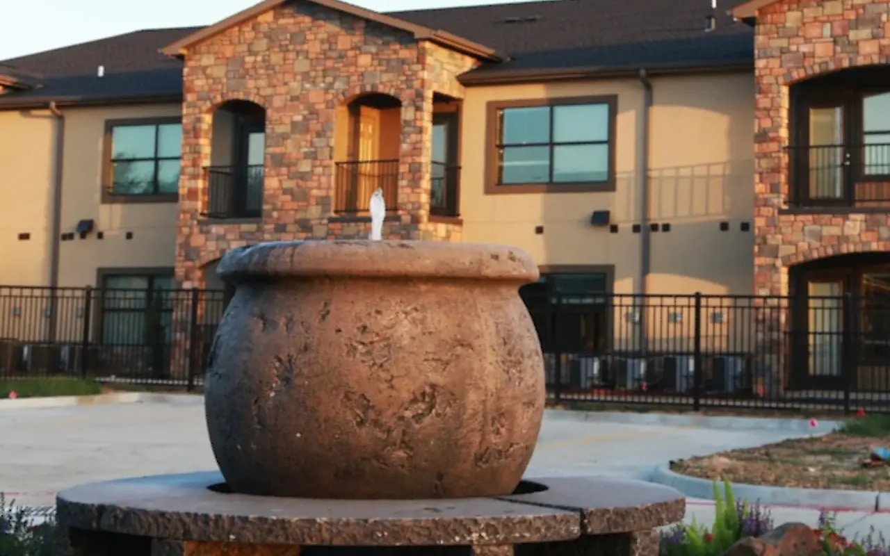A large, round stone fountain set on a stone base, with a trickle of water coming from the top. Behind it, there is a modern residential building with a stone facade and large windows.