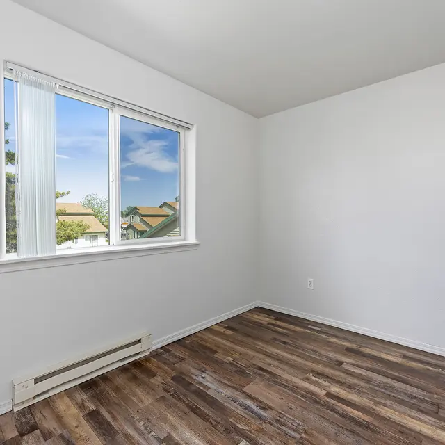Bright Empty Room A vacant room featuring white walls and wooden flooring. A large window allows natural light to illuminate the space, with a view of green trees and houses outside. The room is sparsely decorated, giving it a minimalist appearance.