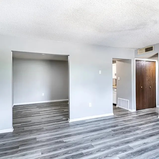 A spacious, bright living room featuring light gray walls and light wood flooring. There are two openings leading to other spaces, one on the left leading to a room and another on the right leading to a kitchen area. The room is well-lit with ample natural light.