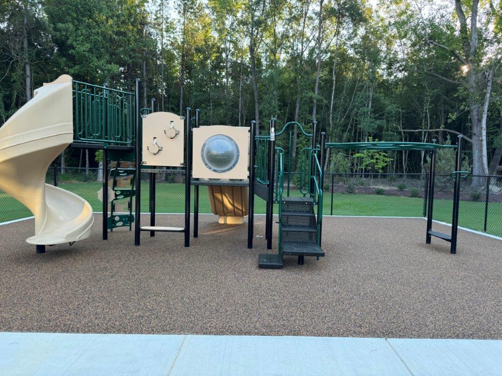 A playground structure featuring a beige slide, multiple climbing areas, and various play elements, surrounded by a grassy area and trees in the background.