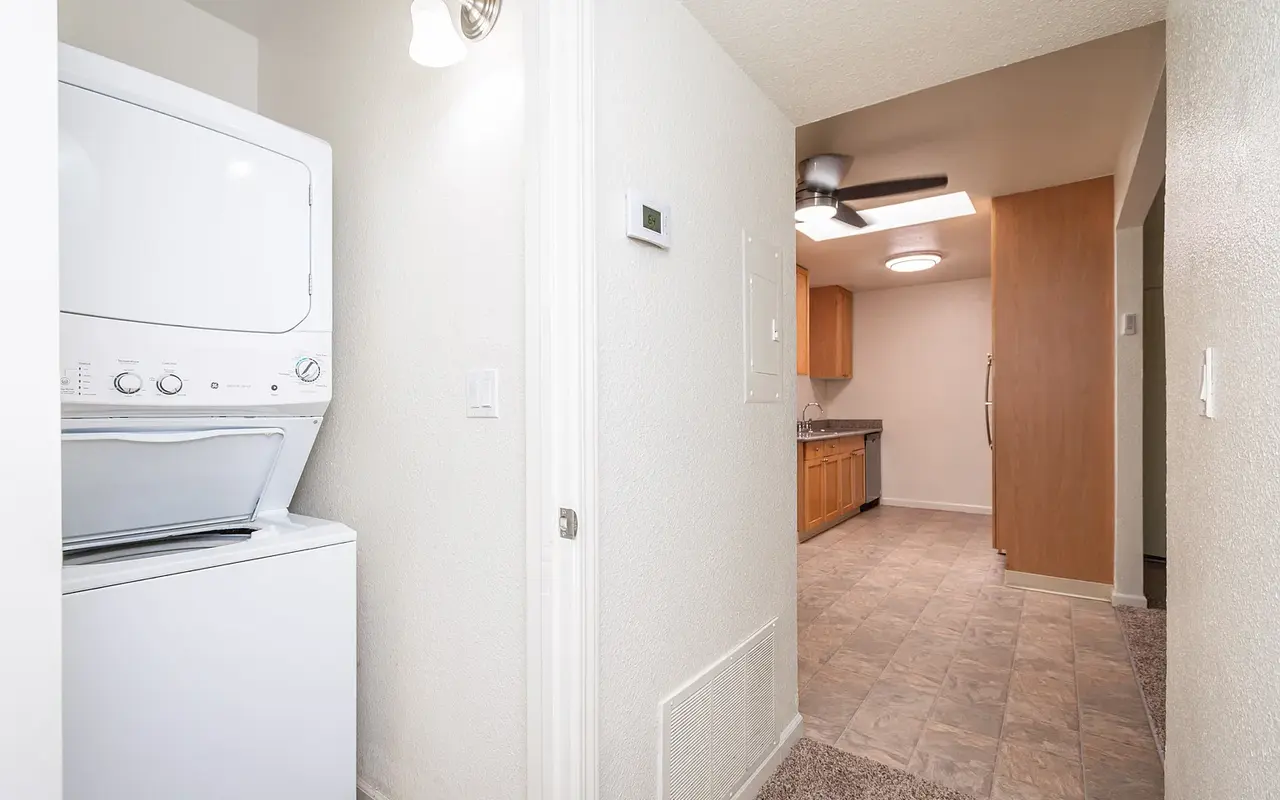 A narrow hallway featuring a stacked washer and dryer on the left, with light-colored walls and a noticeable transition to a kitchen area at the end.