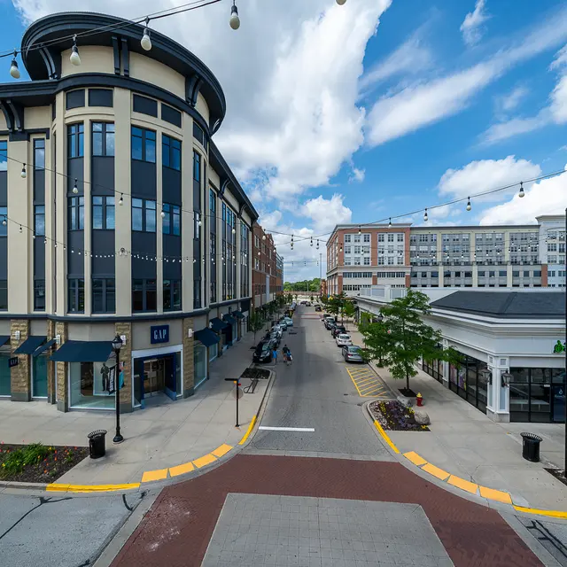 A view of a vibrant urban street lined with buildings, featuring shops and greenery, under a predominantly blue sky with some clouds.