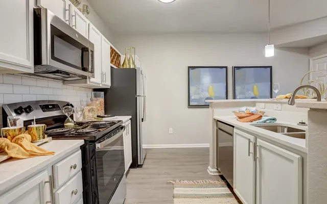 A modern kitchen with white cabinets, stainless steel appliances, and a light-colored countertop. The kitchen features a gas stove, a sleek black refrigerator, and artwork on the walls. A kitchen island with bar seating is visible, along with decorative items and towels on the counter.