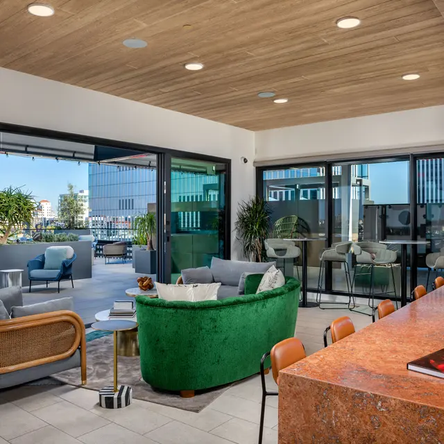 A modern living space featuring a green and gold sofa, gray chairs, and a large kitchen island with an orange countertop. Large windows showcase an outdoor area with plants and city views.