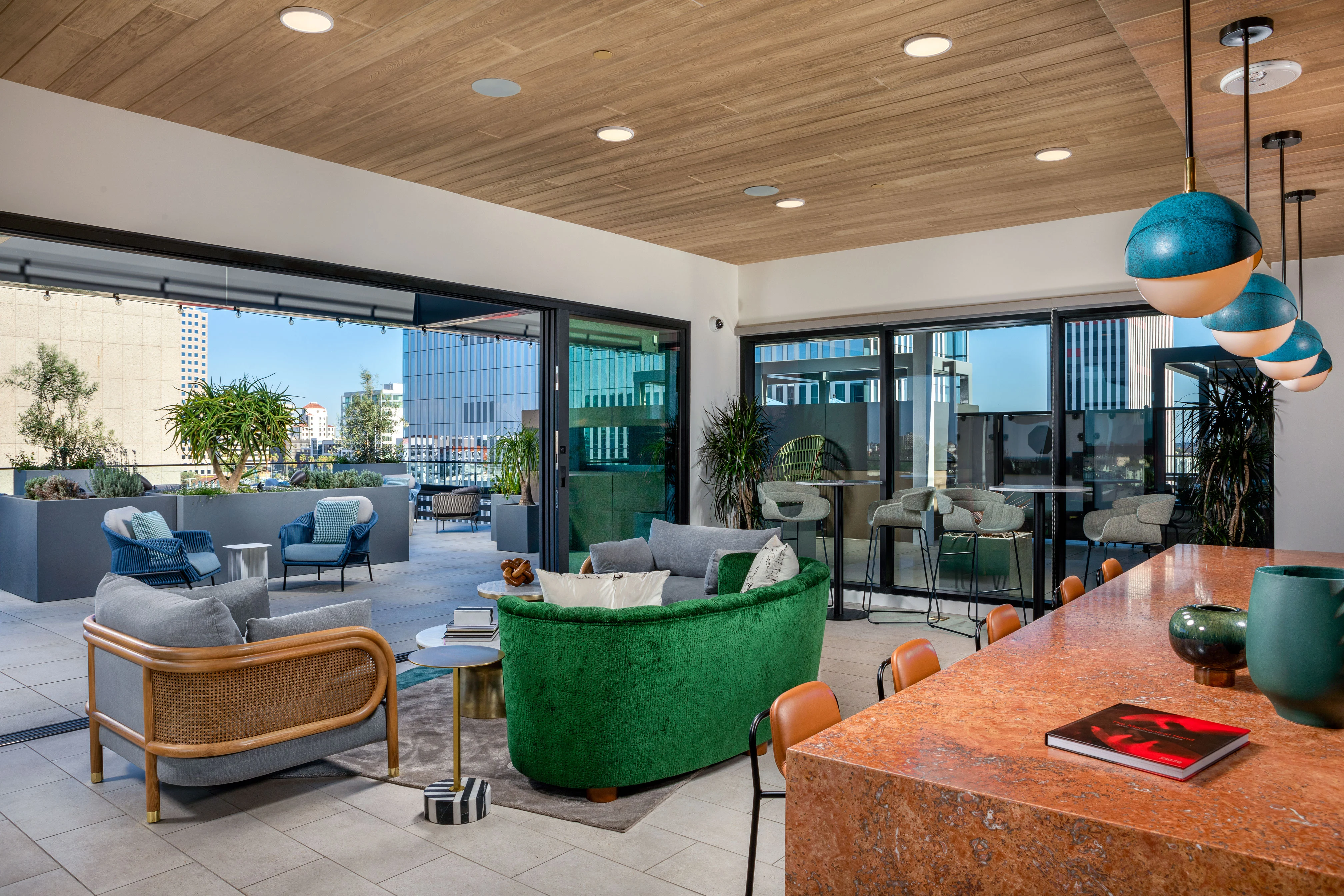 A modern living space featuring a green and gold sofa, gray chairs, and a large kitchen island with an orange countertop. Large windows showcase an outdoor area with plants and city views.