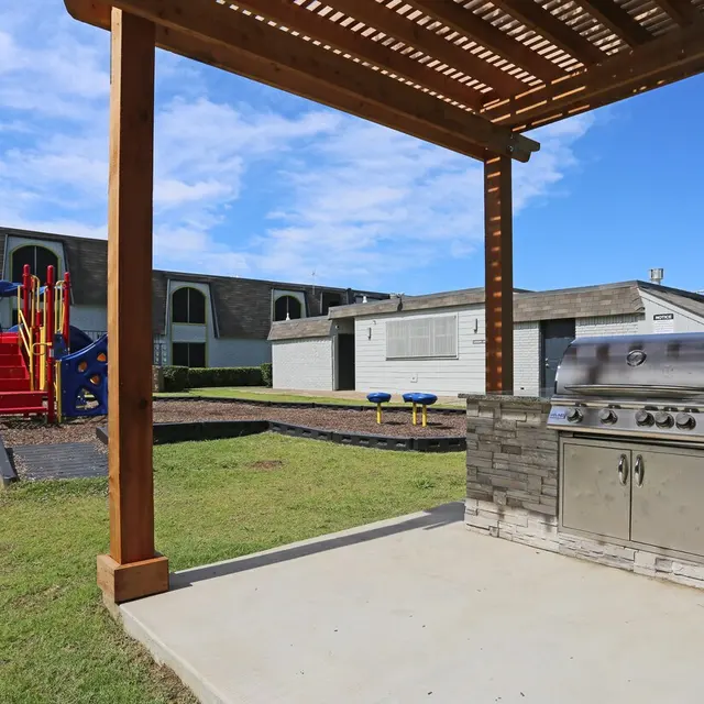 An outdoor barbecue grill area under a wooden pergola with a children's playground visible in the background.