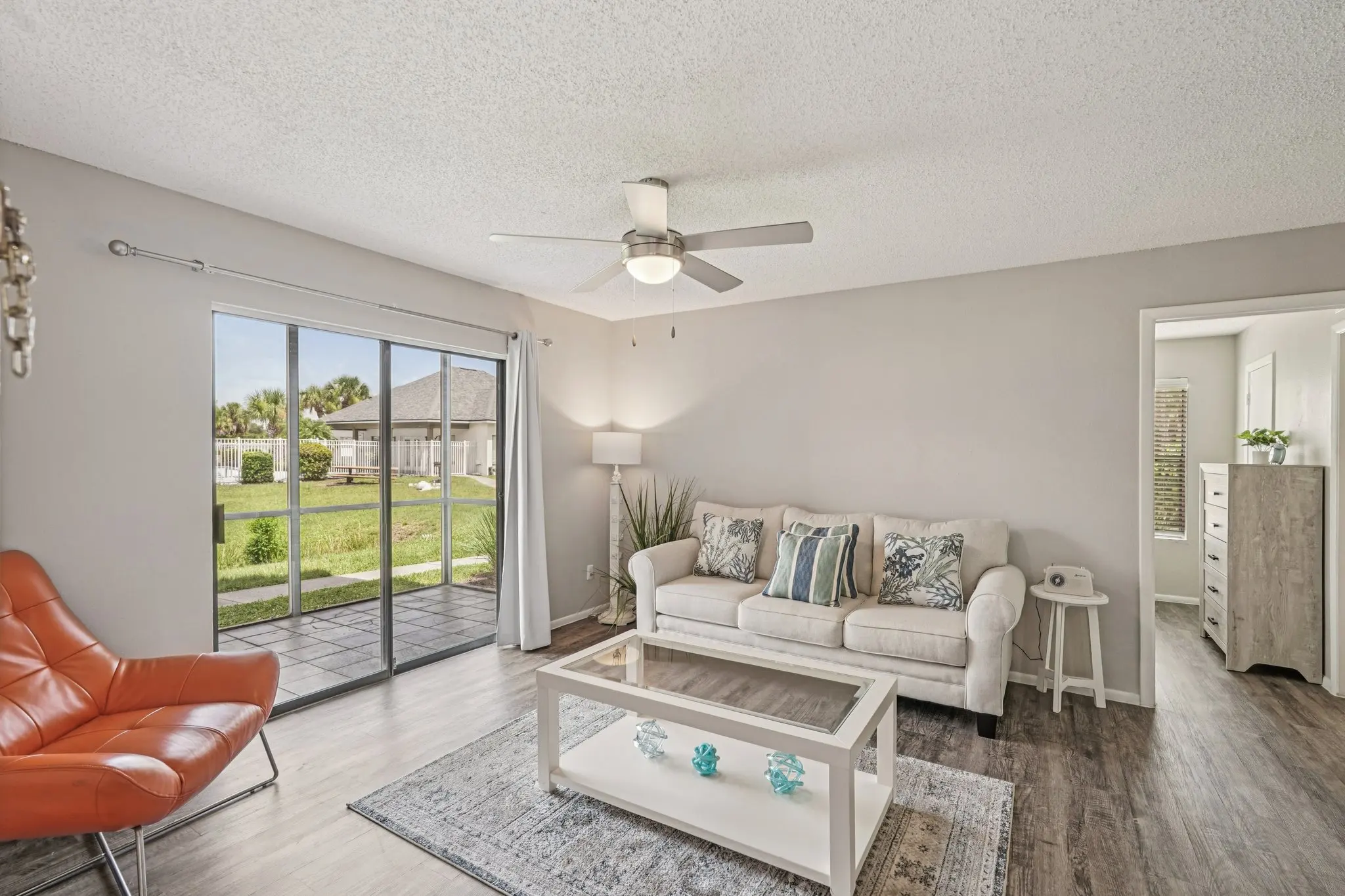 A bright and airy living room with a modern design featuring a cream-colored sofa adorned with blue and white cushions, an orange armchair, a glass coffee table with decorative items, and sliding glass doors leading to a green outdoor view.