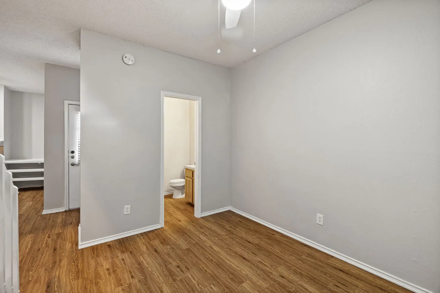 Empty Room with Bathroom Access An empty room with wood laminate flooring and light gray walls, featuring a ceiling fan and a doorway leading to a bathroom.