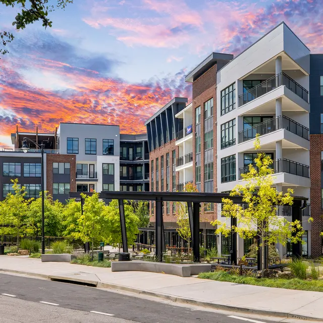 A contemporary apartment complex with a variety of architectural styles surrounded by greenery and trees, set against a colorful sunset sky.
