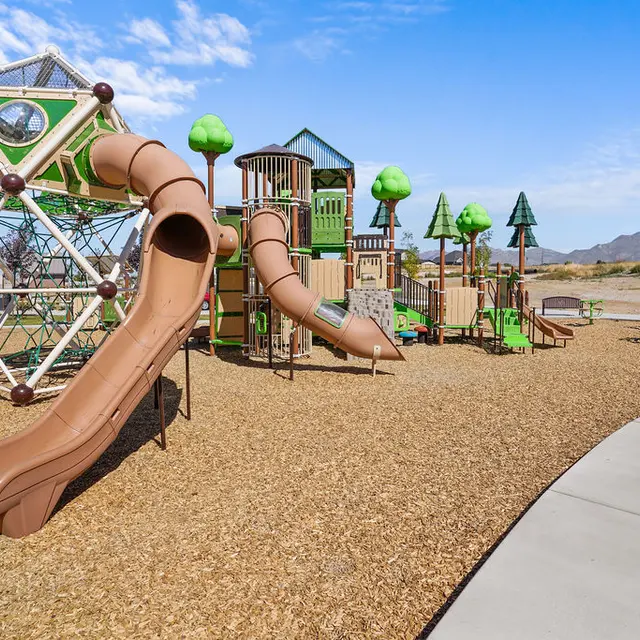A colorful playground featuring various structures, including slides, climbing equipment, and trees, situated on a gravel surface with a clear blue sky.