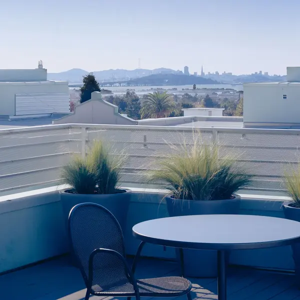 A rooftop terrace with a round table and chairs, surrounded by potted plants. In the background, a city skyline and distant hills are visible under a clear blue sky.