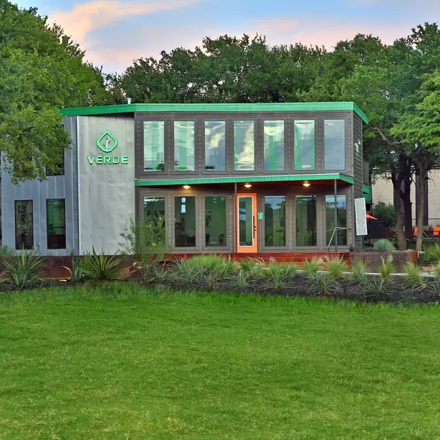 A modern building with large windows and a green roof, featuring the word 'VERDE' prominently displayed. The surroundings are landscaped with green grass and trees. The setting suggests a serene environment with a cloudy sky at dusk.