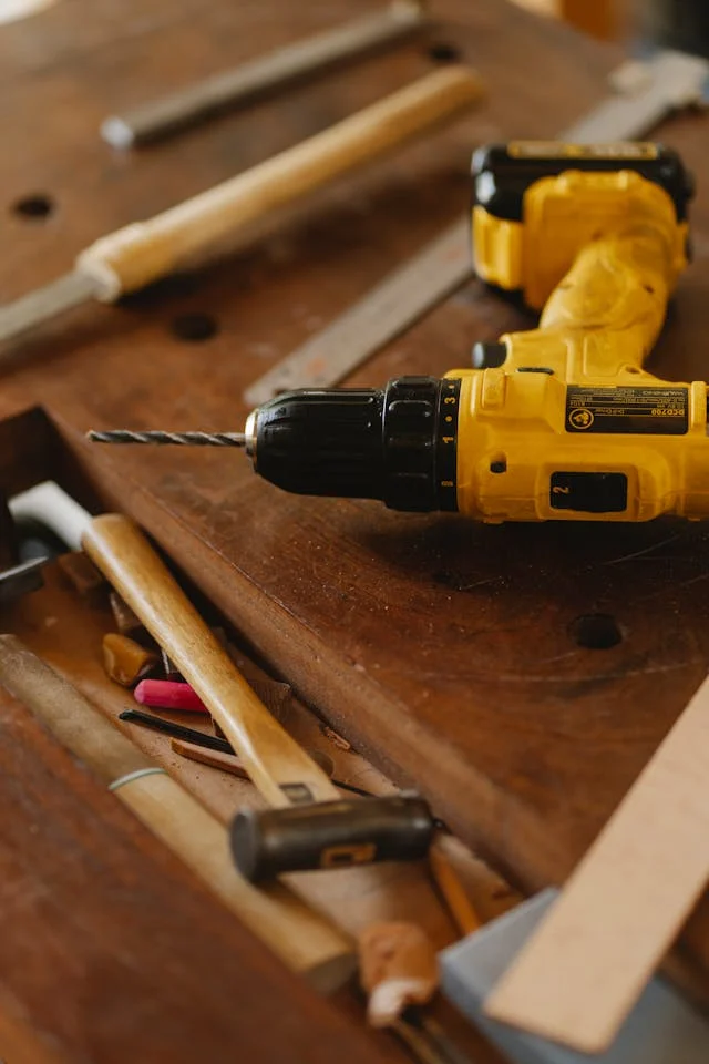 A yellow handheld drill rests on a wooden workbench surrounded by various woodworking tools and materials.