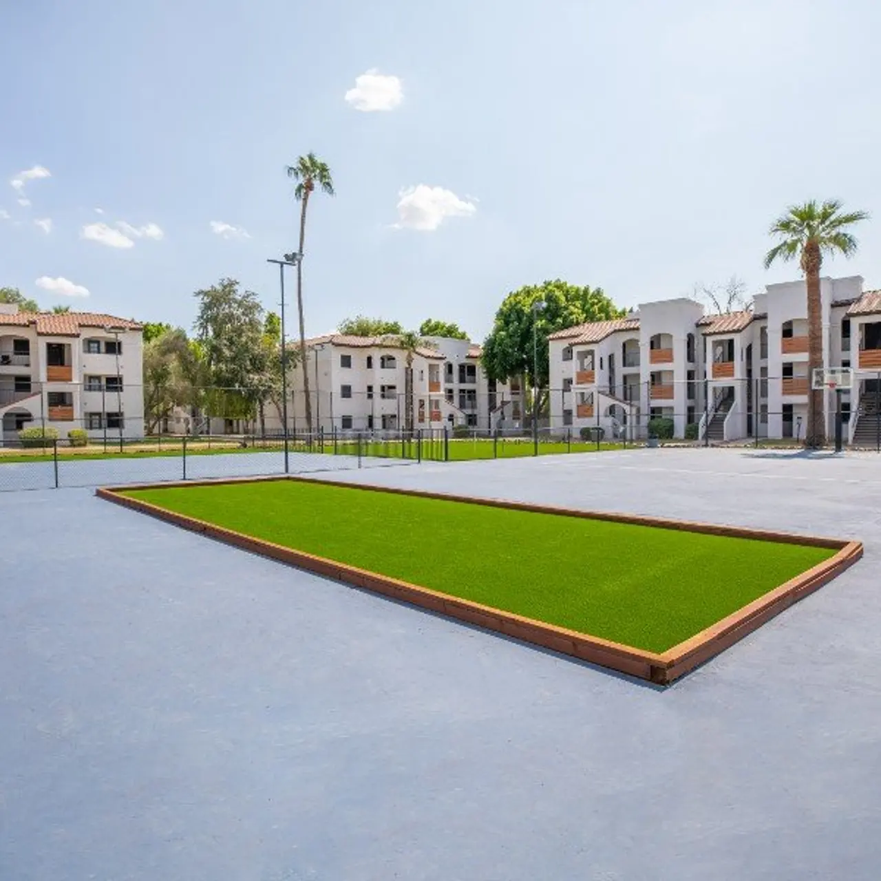 A spacious courtyard in an apartment complex featuring a small green lawn area surrounded by a concrete surface and palm trees, with multiple apartment buildings in the background.