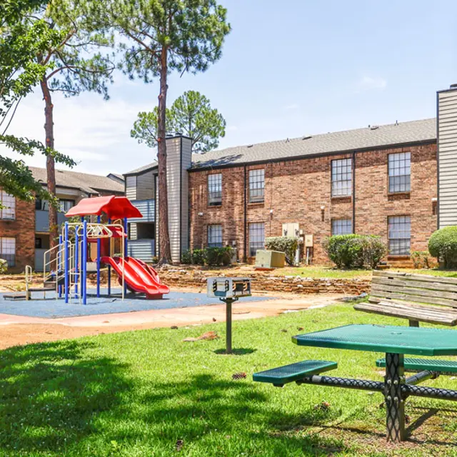 A playground with red slides and climbing structure, surrounded by green grass, trees, and brick apartment buildings in the background.