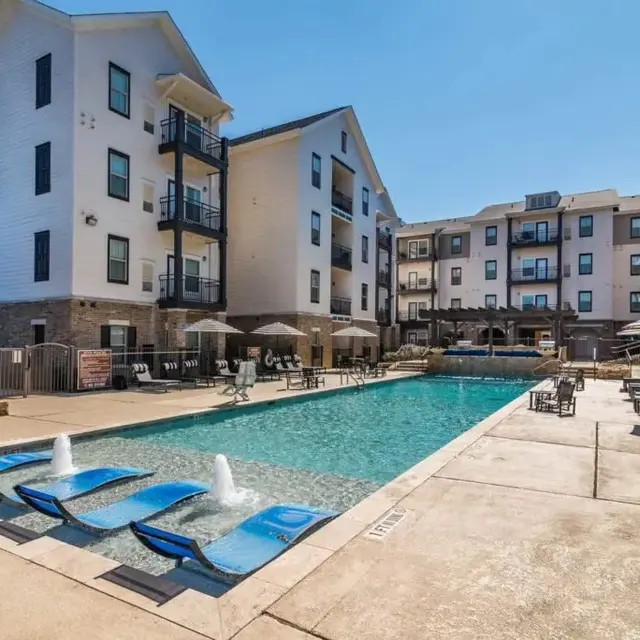 A modern swimming pool surrounded by apartment buildings, featuring sun loungers and umbrellas.