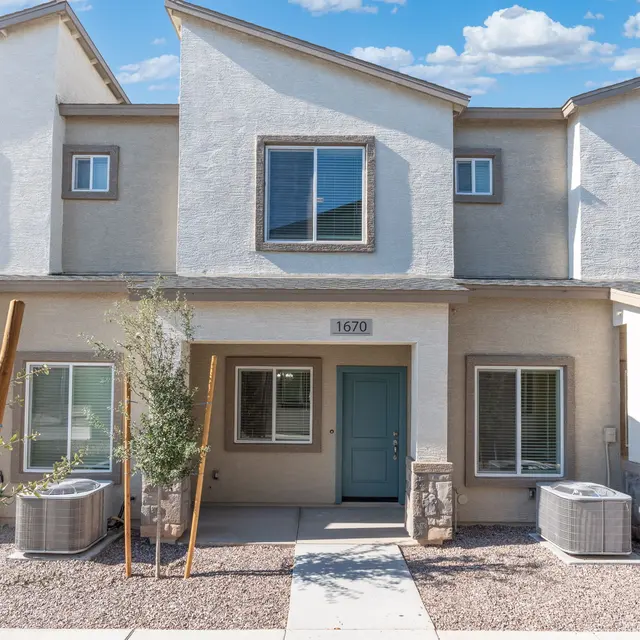 Modern Townhouse Exterior A row of newly constructed townhouses featuring a central entrance with a blue door, flanked by two air conditioning units and small shrubs in front.