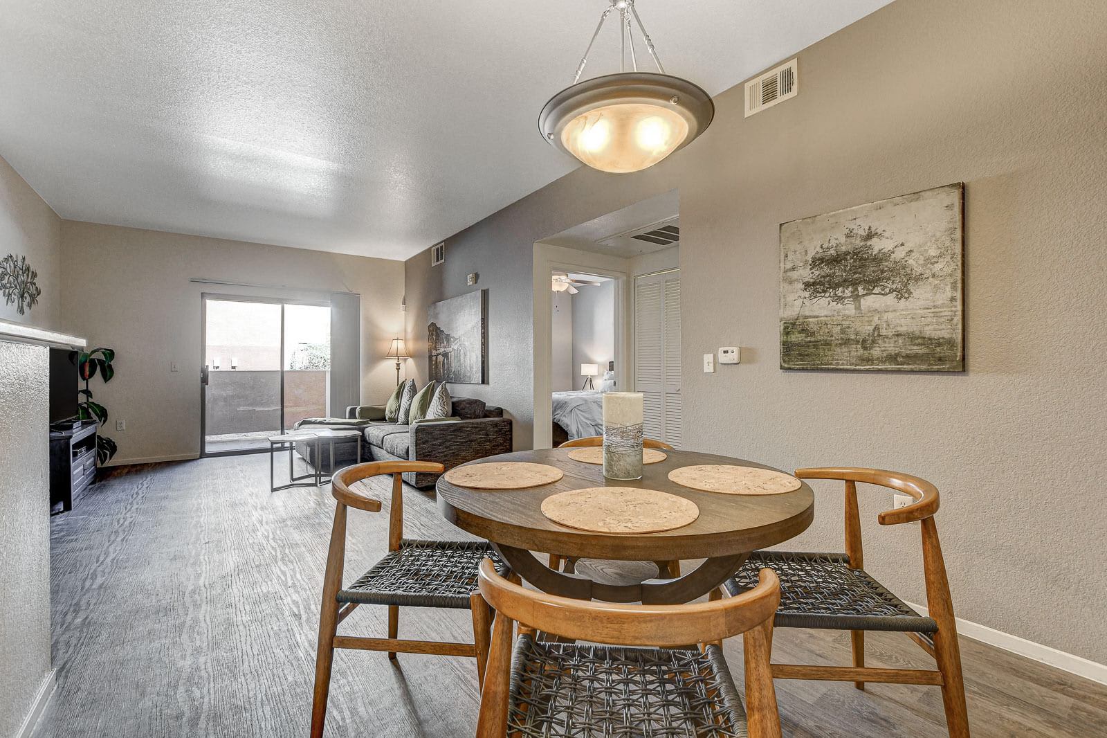 Chic Dining Area in a Contemporary Apartment A modern dining area featuring a circular table with place settings, surrounded by wooden chairs. In the background, a cozy living space with a couch and sliding glass door leading to an outdoor area can be seen.