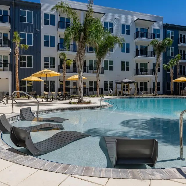 A modern apartment complex with a swimming pool, lounge chairs, and umbrellas under a clear blue sky.