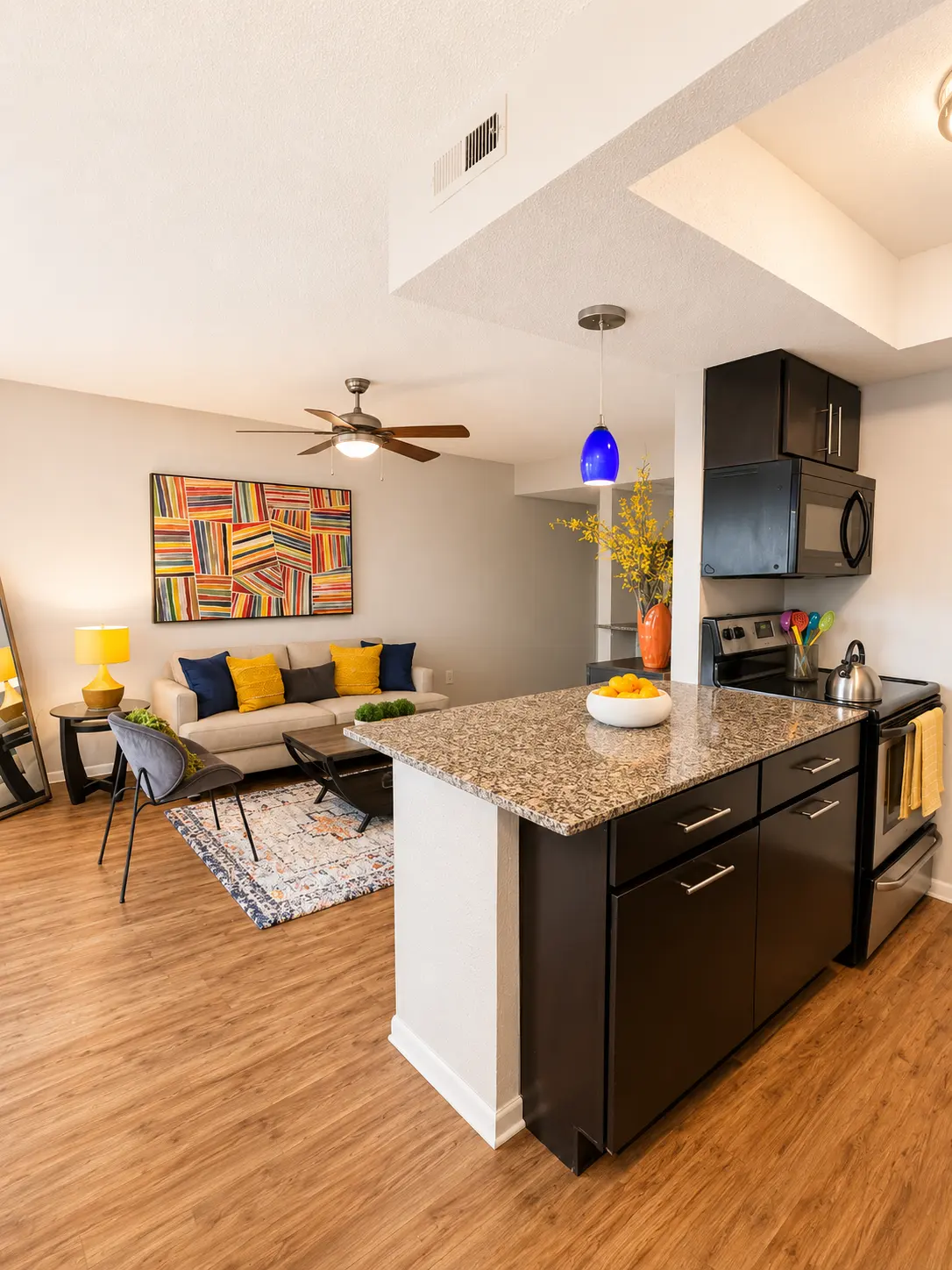 A modern apartment interior featuring an open kitchen and living area. The kitchen has dark cabinetry and granite countertops, with a stainless steel stove. A colorful painting hangs on the wall of the living area, which includes a light gray sofa adorned with yellow and blue pillows, and a decorative coffee table on a patterned rug.