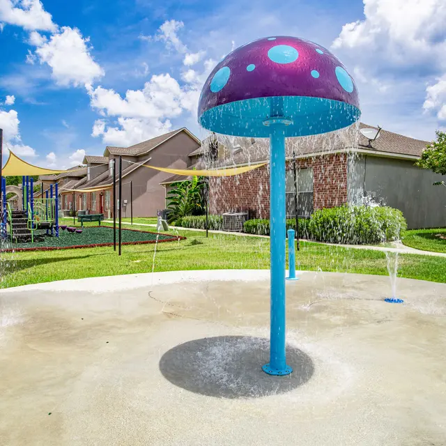 A colorful water spray feature in a playground area, resembling a mushroom with blue and purple colors. Surrounding it are green lawns and a playground set in the background under a bright blue sky with fluffy clouds.