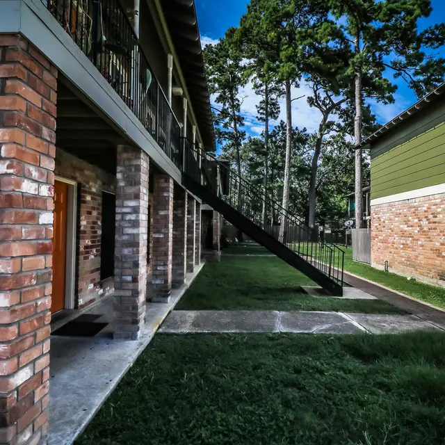 Exterior view of an apartment complex featuring brick columns, a stairway, and green trees in the background.