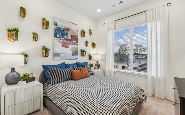 A modern bedroom featuring a bed with black and white striped bedding and decorative pillows. The walls are adorned with planters and a colorful abstract painting. Natural light enters through a window covered by sheer curtains. There's a nightstand with a lamp and a bedside plant.