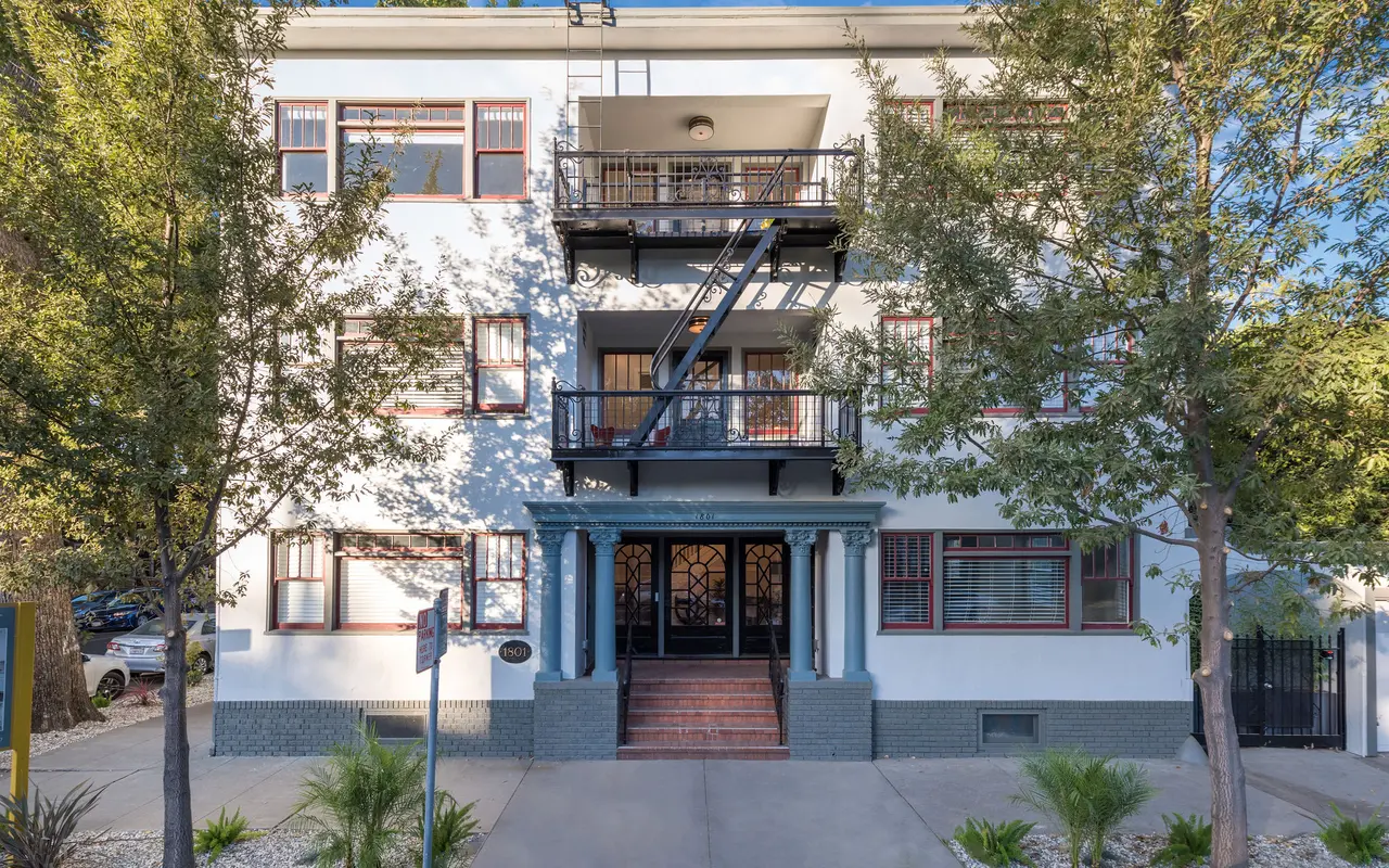 Front view of a three-story apartment building with green trim, balconies, and surrounding trees.