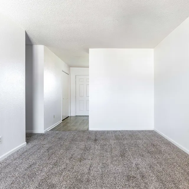 Empty living room with carpet and white walls, featuring a closed door and a small hallway