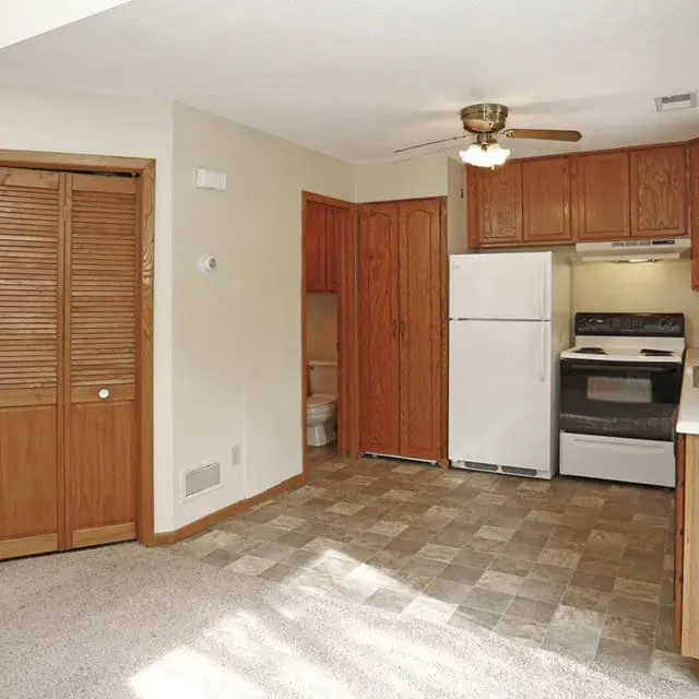 Compact Kitchen Interior A small kitchen area with wooden cabinets, a white fridge, and a stove, featuring carpeted flooring and a ceiling fan.