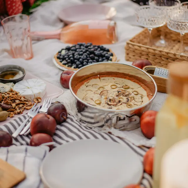 A beautifully arranged picnic spread on a blanket, featuring various dishes and drinks. There are plates with cheese, olives, nuts, and fruits, alongside a bowl of dessert or dip. A basket and some wine glasses are visible in the background, creating a cozy atmosphere.