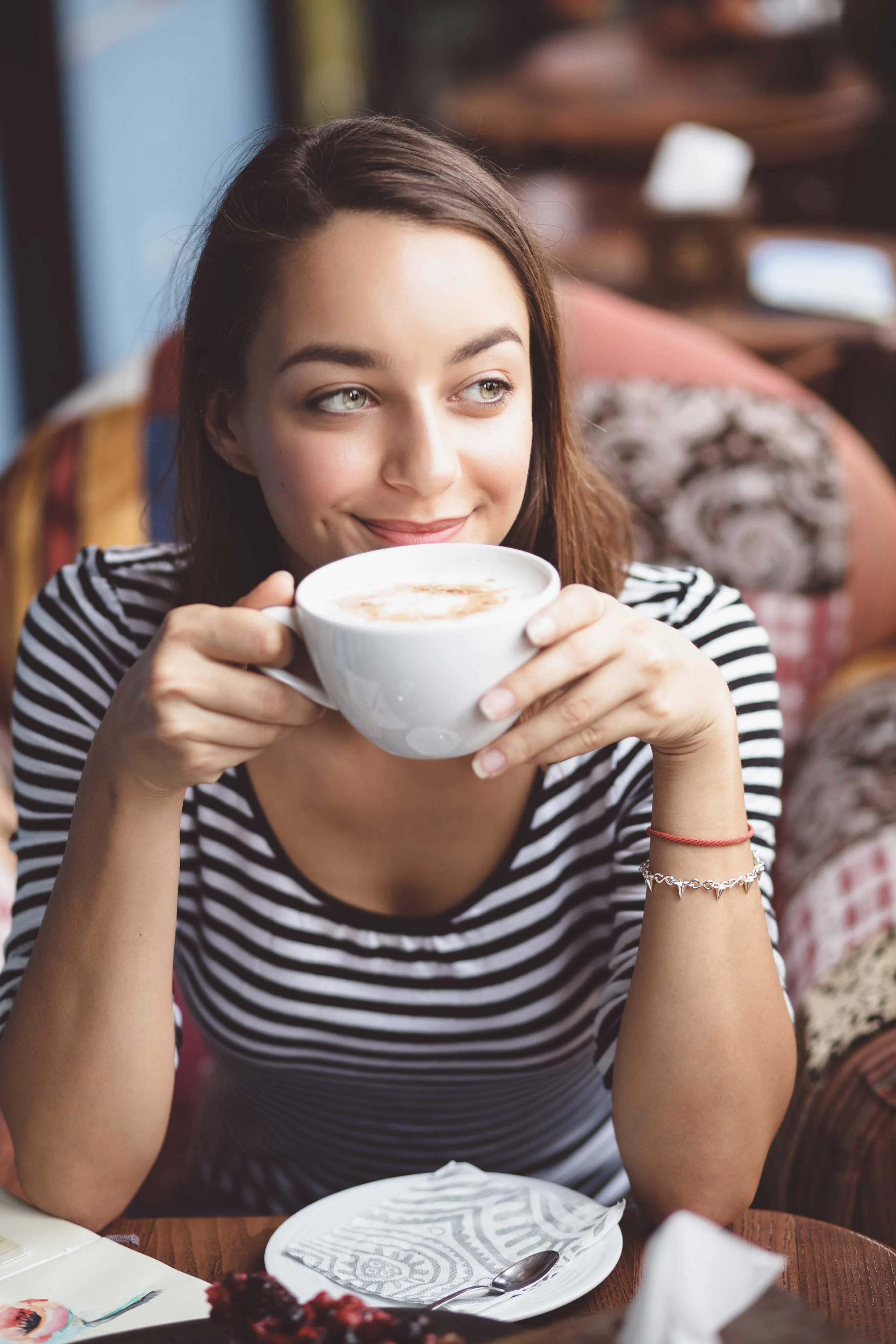 A young woman sitting leisurely in a cozy café, holding a large cup of coffee with a smile, wearing a striped shirt.