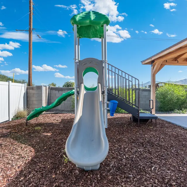 A playground slide with a green top and gray body, surrounded by wood chips and a white fence, with trees and a pavilion in the background under a blue sky with clouds.
