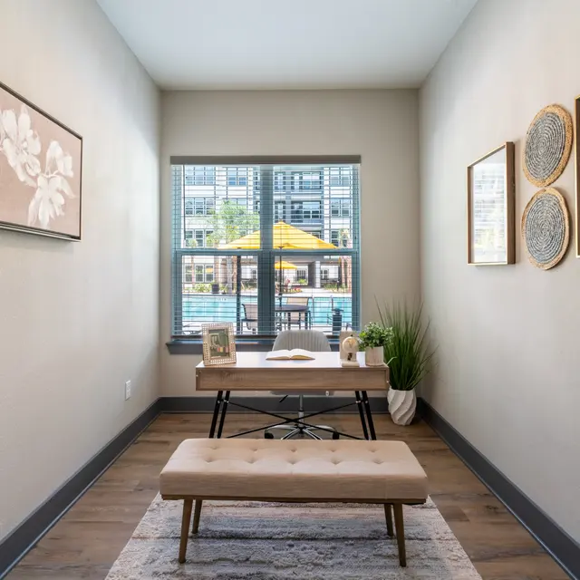 Modern Workspace with Outdoor View A modern workspace featuring a wooden desk, a small bench, and framed wall art, with large windows showing an outdoor view.