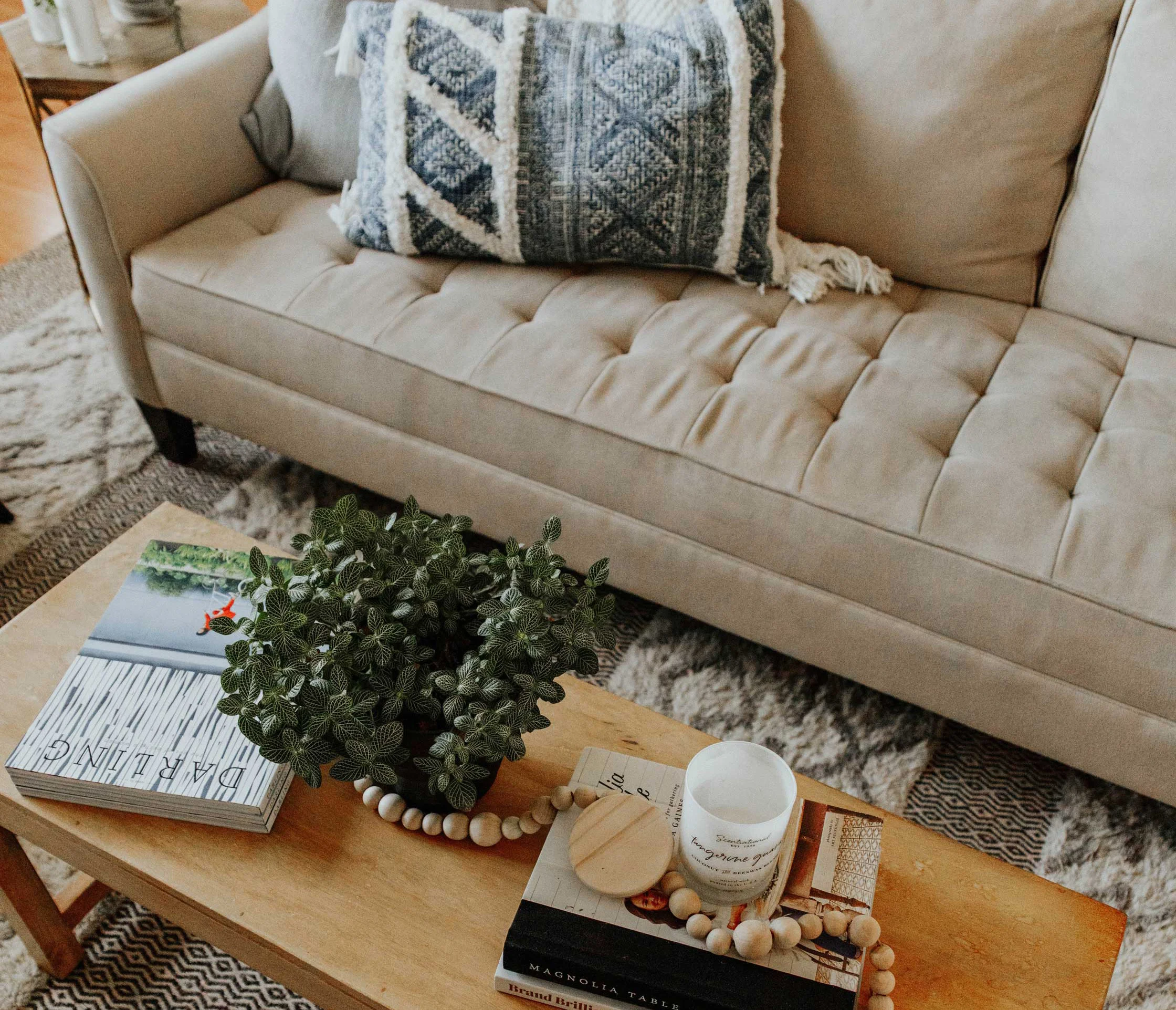 Cozy Living Room Setup A cozy living room with a beige sofa adorned with decorative pillows, a wooden coffee table topped with books, a candle, and a small potted plant.