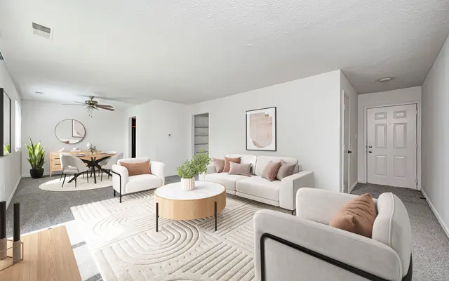 Contemporary Living Room Design A modern living room featuring white walls, soft carpets, and contemporary furniture. There are two white sofas with decorative pillows, a round coffee table in the center, and a dining area in the background with a round table and chairs. A plant adds a touch of greenery, and there are large windows allowing natural light to brighten the space.