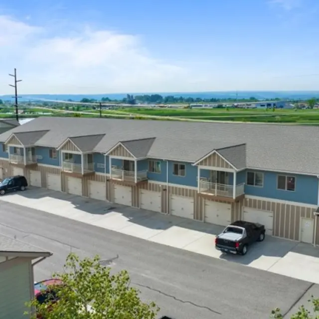 Granite Peak Aerial view of a multi-unit residential building featuring several blue and beige units with porches and garages. A parking area is visible in front with several cars parked. In the background, green fields and a clear blue sky can be seen.