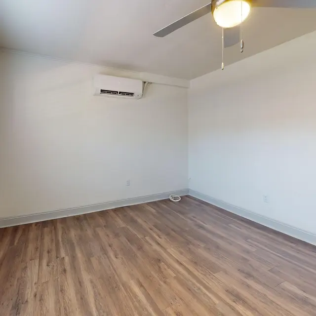 A vacant living room with light-colored walls and wooden flooring. There is a ceiling fan and an air conditioning unit visible. A window with blinds lets in natural light.