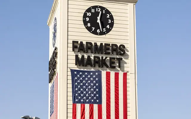 A tall clock tower displaying the words 'FARMERS MARKET' with an American flag draped below. The sky is clear and blue, and there are signs and a building below the tower.