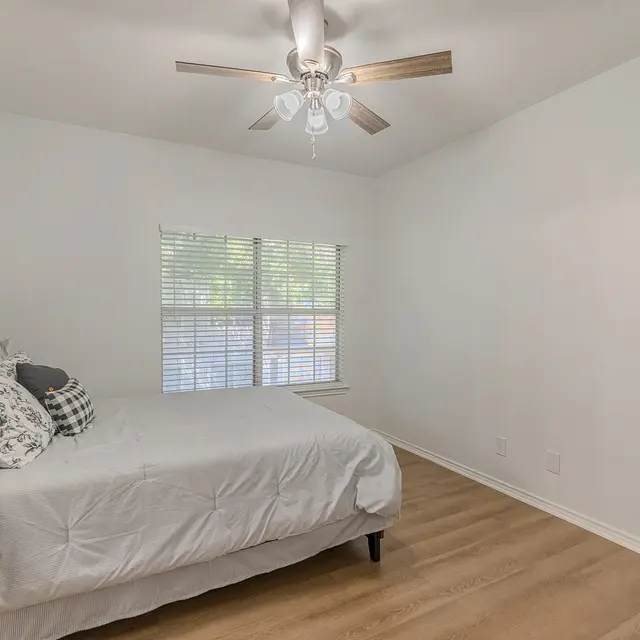 A spacious and bright bedroom featuring a queen-sized bed with white linens and decorative pillows, a wooden nightstand, and a ceiling fan. The room has white walls and wooden flooring, with a window that has blinds allowing natural light to enter.