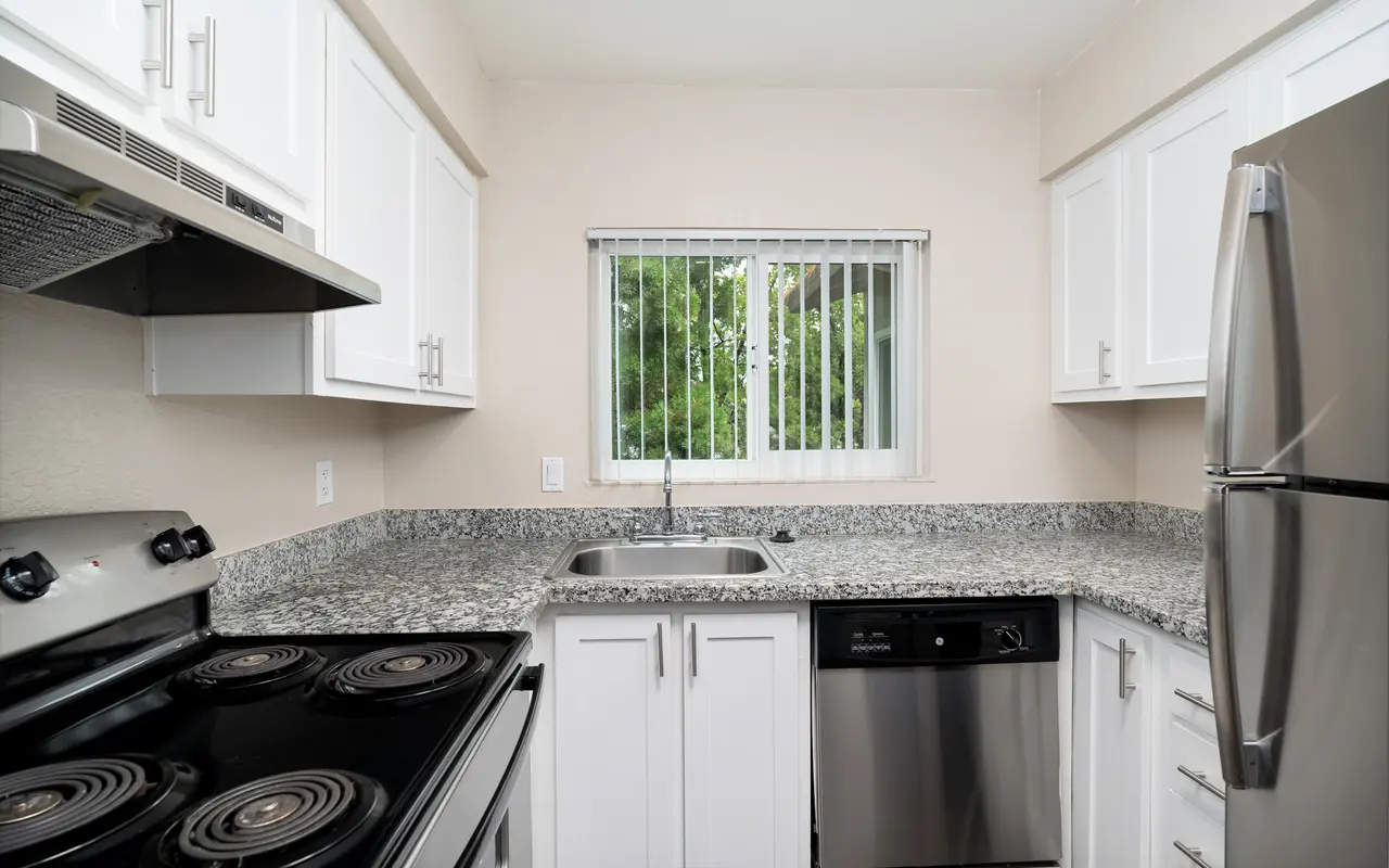 A modern kitchen featuring white cabinets, stainless steel appliances, and a granite countertop. Includes a stove, refrigerator, and dishwasher with a window overlooking greenery.