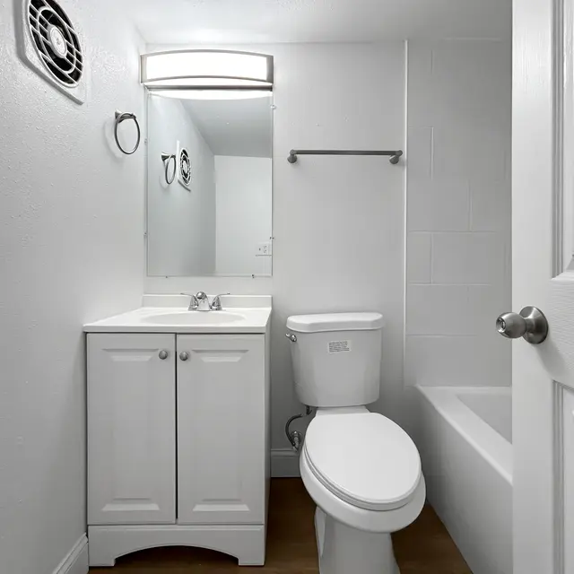 Modern Bathroom Interior A clean and modern bathroom featuring a white vanity with a sink, a toilet, and a bathtub. The walls are painted white, and there's a mirror above the sink with a light fixture. A small vent is visible on the ceiling.
