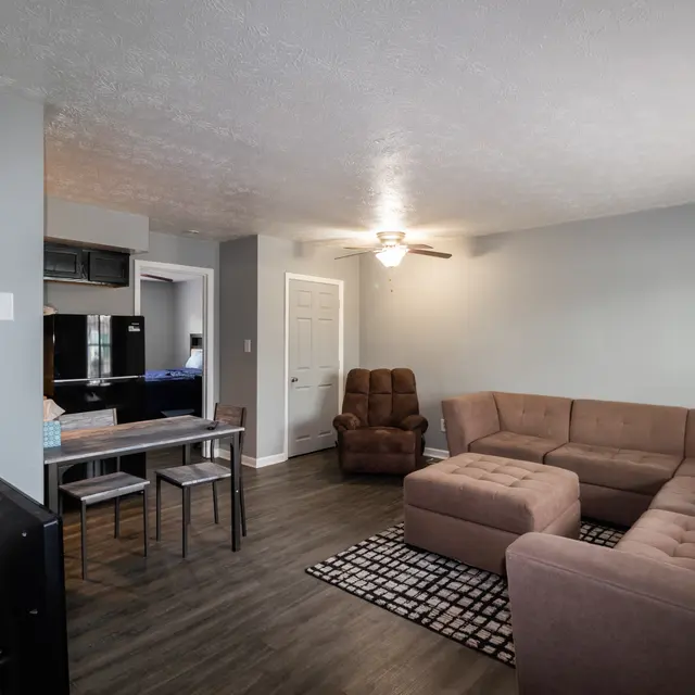 A living room featuring a light gray wall, a large brown sectional sofa, a black and white patterned rug, a round dining table with four chairs, a recliner, and kitchen access in the background.