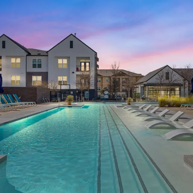A beautifully designed swimming pool area at a modern apartment complex during dusk. The pool features lounge chairs and a serene atmosphere with a colorful sky in the background.