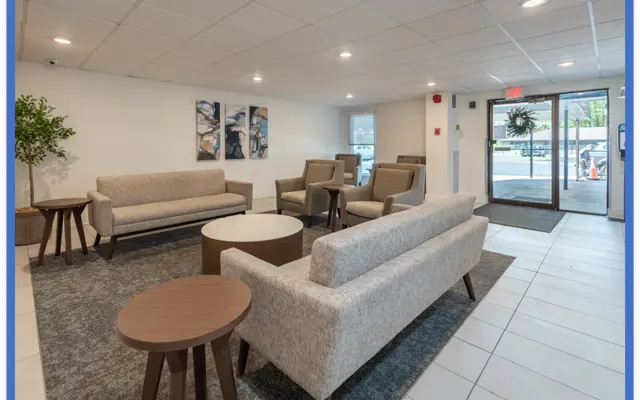 A modern lobby area featuring gray upholstered sofas and round tables on a dark rug, with artwork on the wall and large windows allowing natural light.
