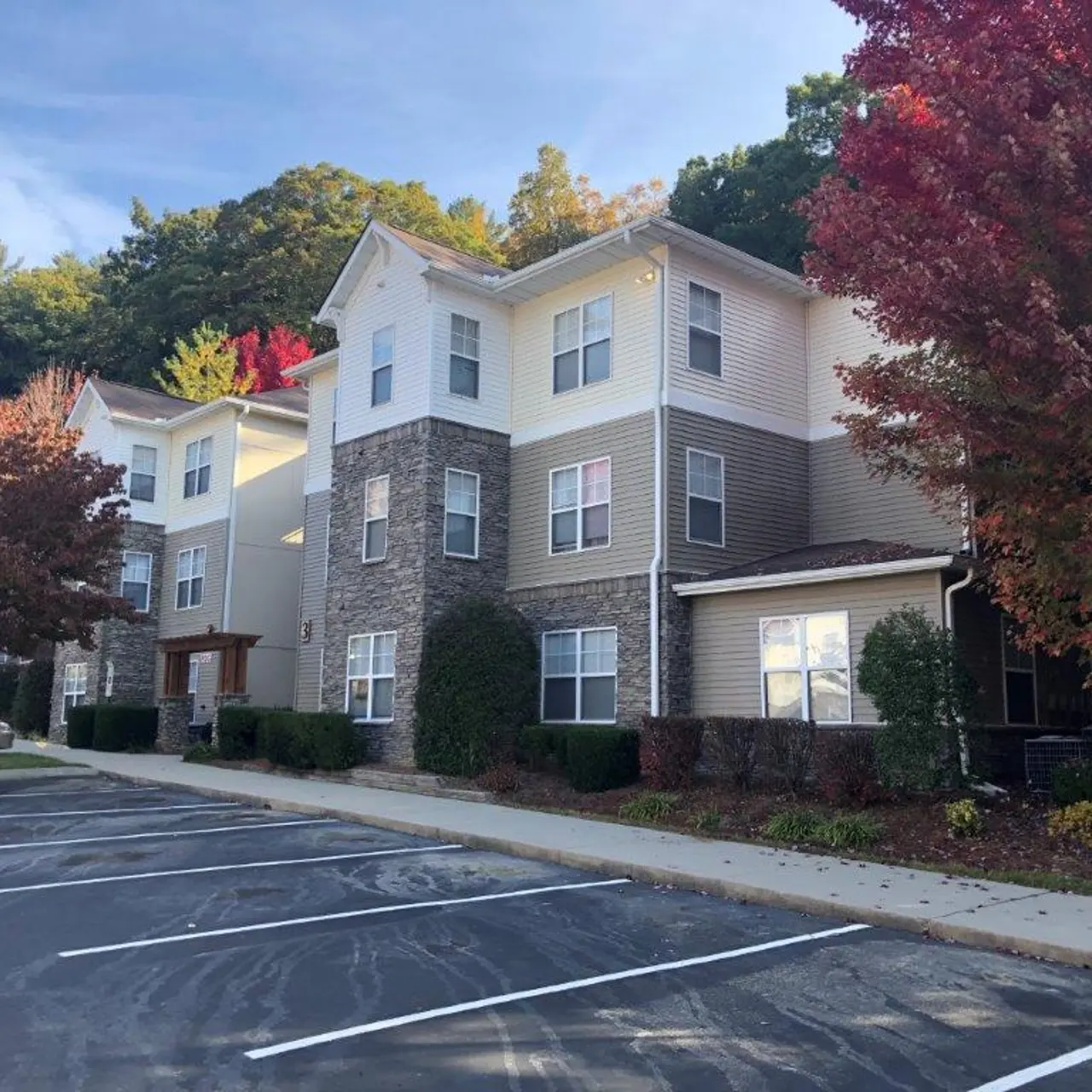 A modern apartment building featuring a mix of stone and siding, with landscaped shrubs and colorful autumn trees in the foreground.