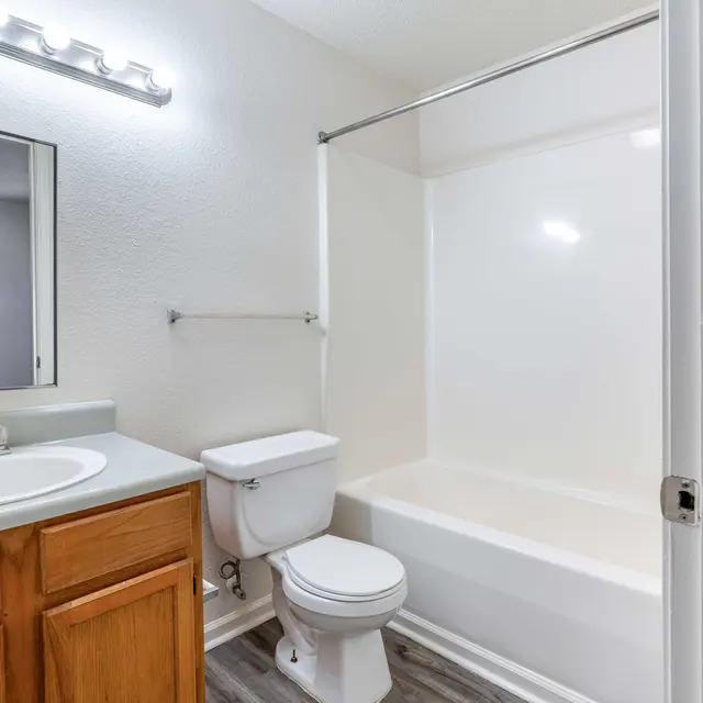 A modern bathroom featuring a white bathtub, a toilet, and a sink with wooden cabinets. The bathroom is well-lit with a light fixture above the mirror.