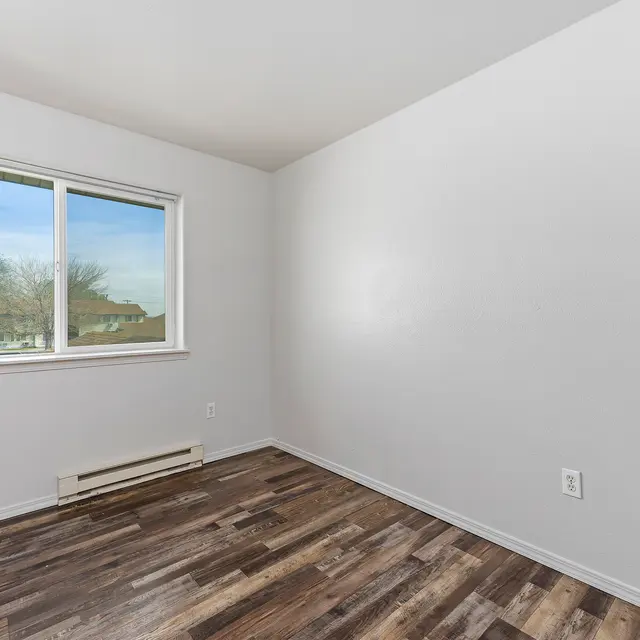 Vacant Room Interior A vacant room with light-colored walls and wooden flooring, featuring a large window that lets in natural light. The room is empty with no furniture present.